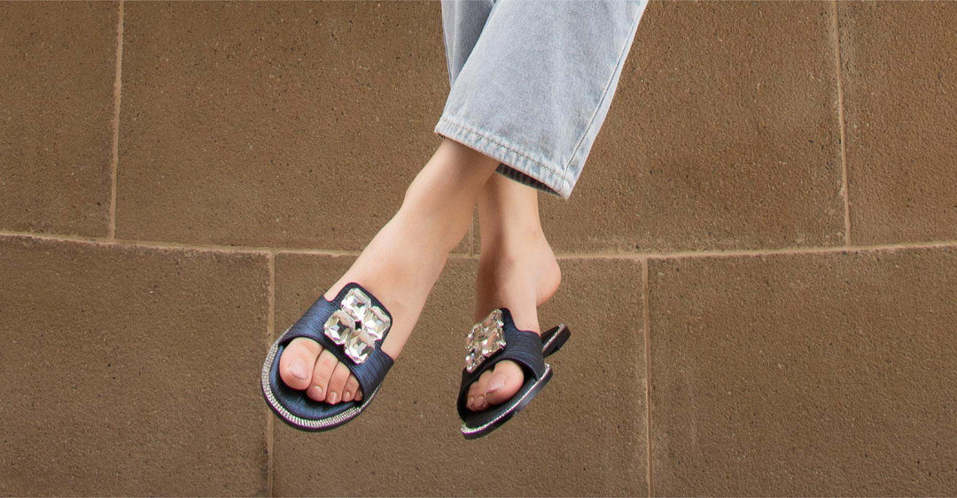 Person wearing stylish black open-toe sandals with decorative buckles, feet dangling against a brown textured wall for online shoe shopping tips.