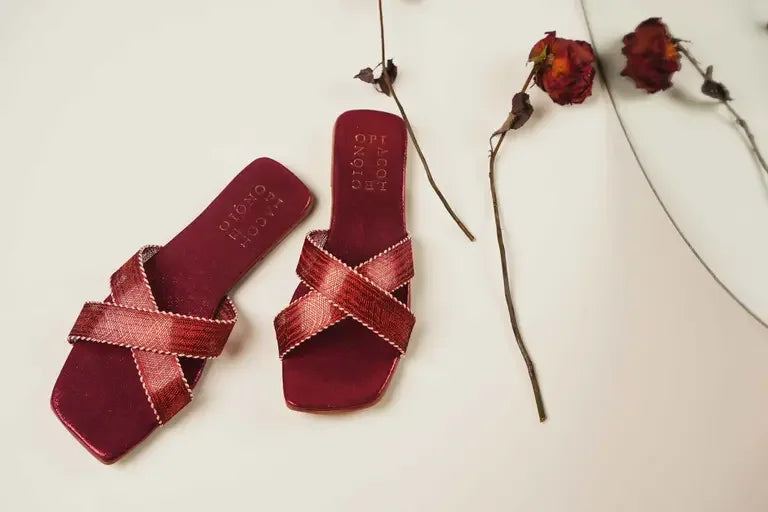 Maroon trendy flat slippers with crisscross straps displayed alongside dried flowers.